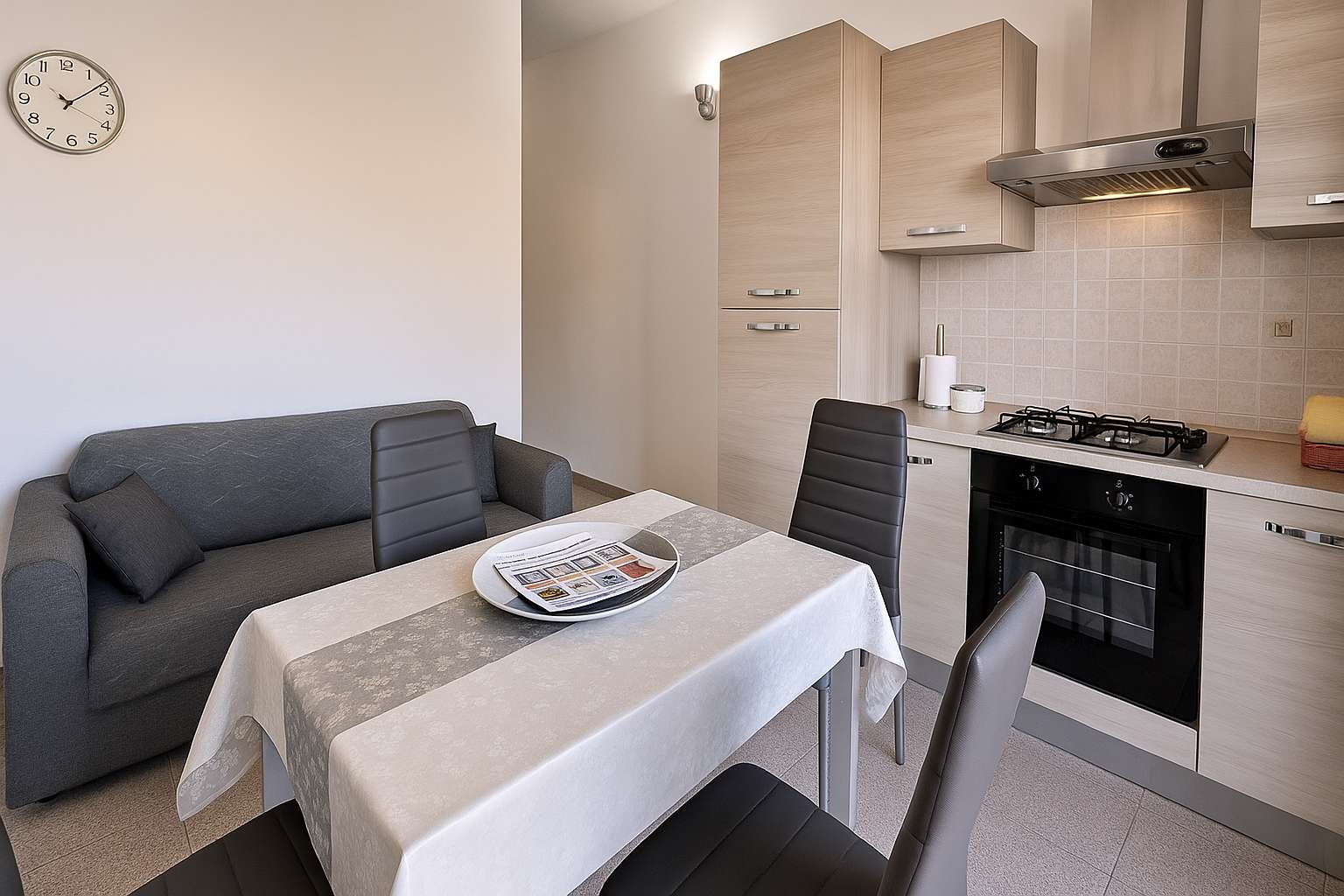 Zona giorno con divano grigio, tavolo apparecchiato e cucina con forno